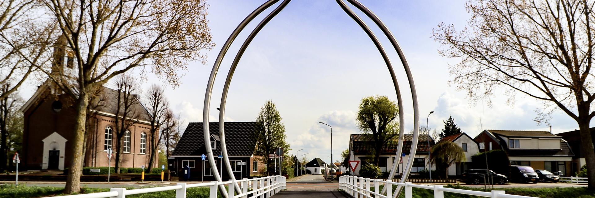 Een sierlijke stalen boog overspant als een welkomstpoort de brug over de Hoofdvaart in Abbenes. Aan weerszijden hangen vlak boven het water twee schalen, als was de constructie een grote weegschaal. De boog wordt bekroond door een metalen bol.